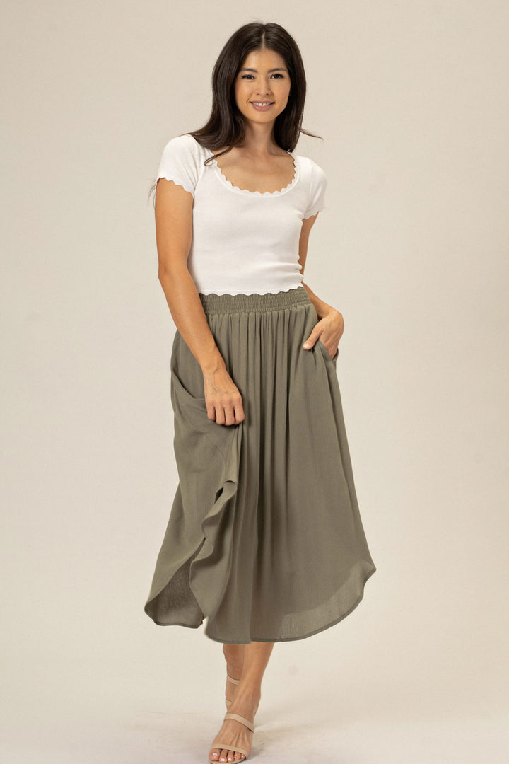 A woman wearing a gray pleated midi skirt with a comfortable elastic waistband, paired with a white scalloped tank top.