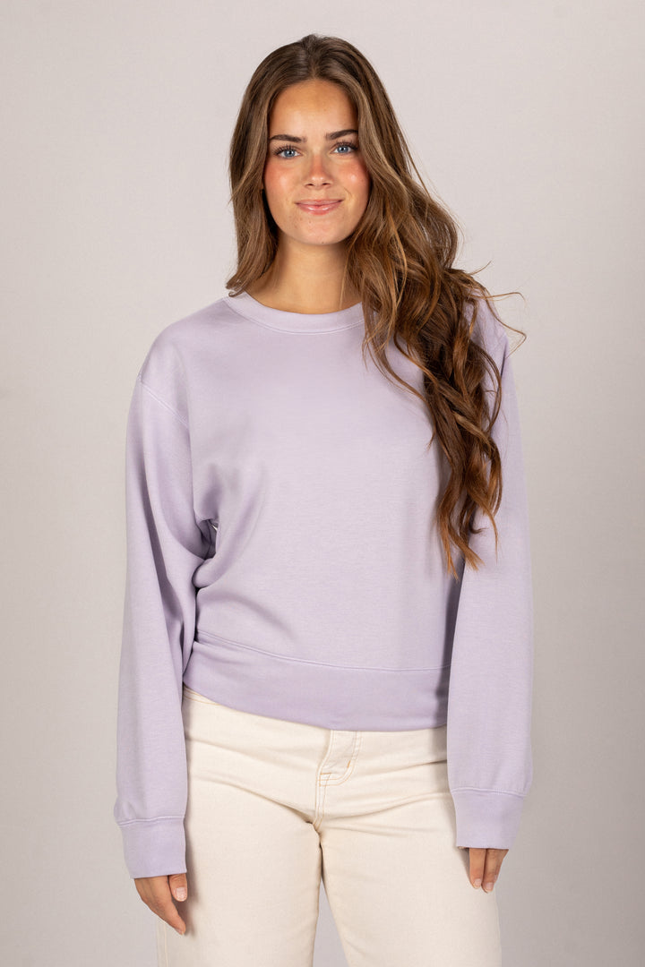 A young woman with long brown hair is wearing a relaxed, light purple sweatshirt with a simple round neckline.