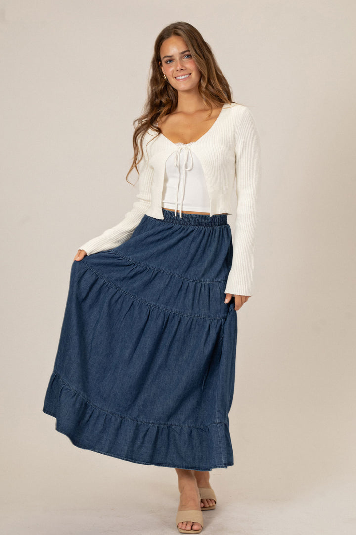 A woman in a white cropped sweater and a long navy blue tiered denim midi skirt, standing against a beige background.