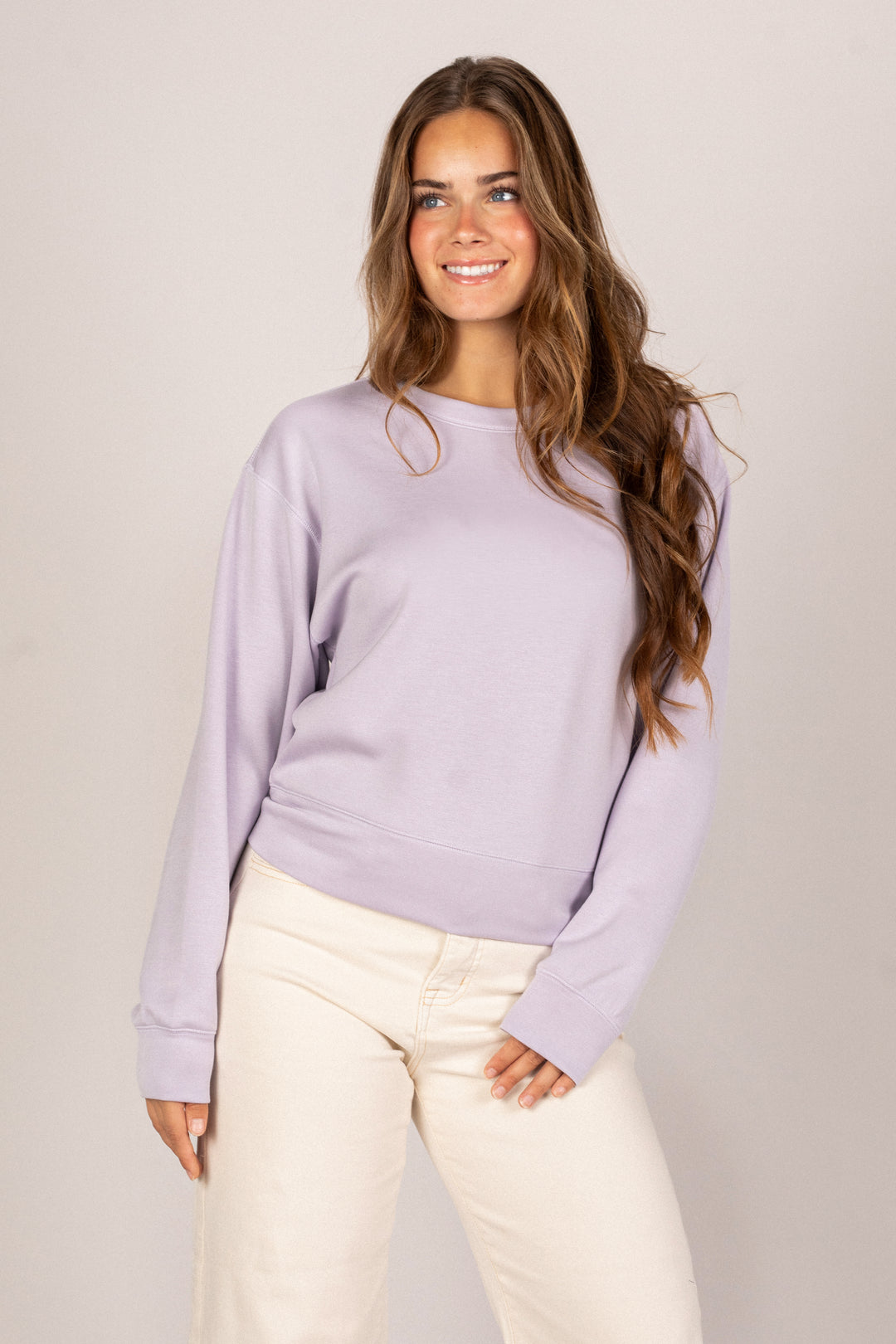 A smiling young woman with long brown hair wearing a relaxed, light purple sweatshirt and off-white pants.