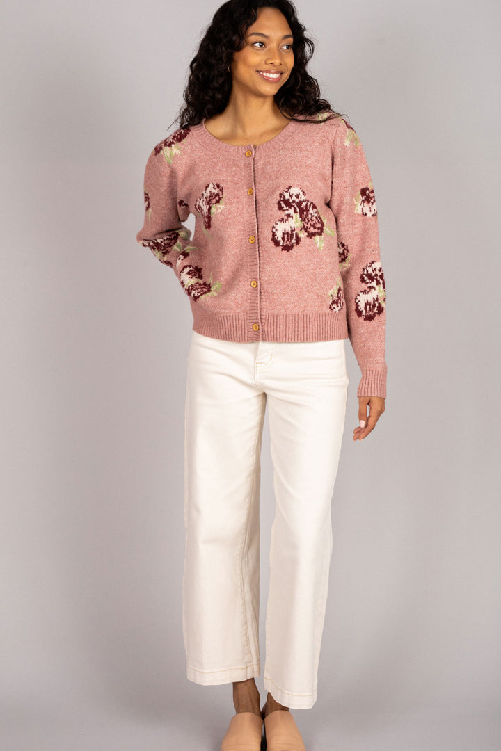A pink and burgundy floral knit cardigan with button-down closure, worn by a smiling woman with curly dark hair in white pants.