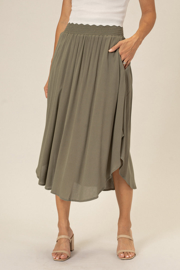 A flowing olive green midi skirt with an elastic waistband and a scalloped hem, worn with nude platform sandals.