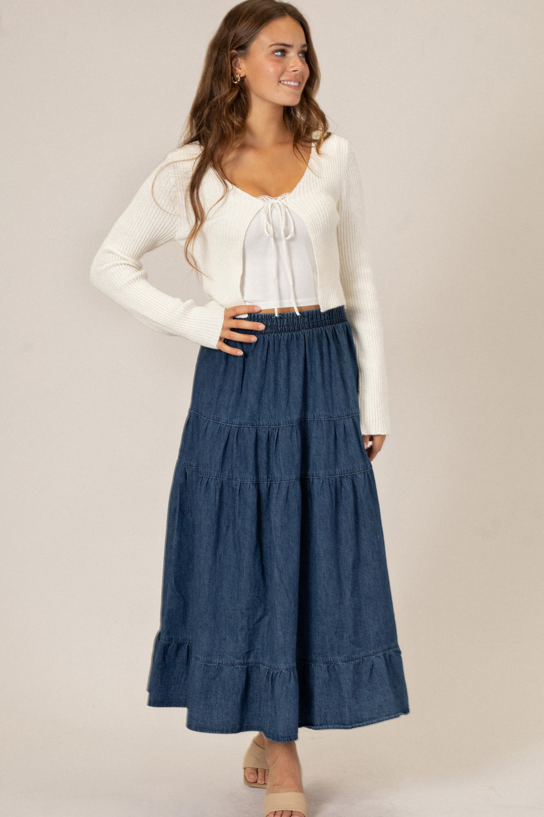 A young woman wearing a white sweater and flowing denim midi skirt with a smocked waistband and tiered ruffled design.