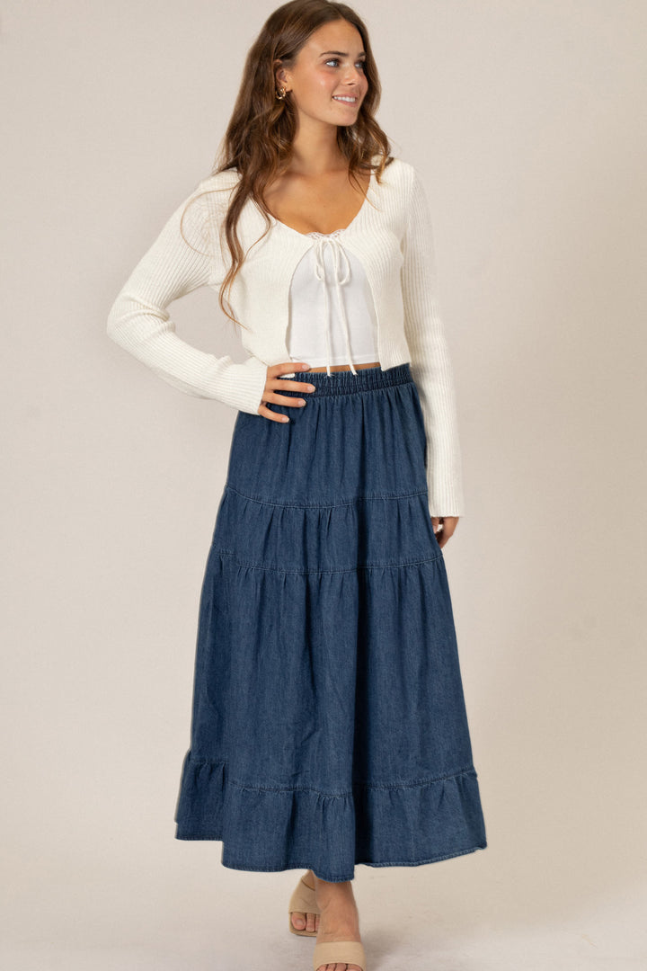 A young woman wearing a white sweater and flowing denim midi skirt with a smocked waistband and tiered ruffled design.