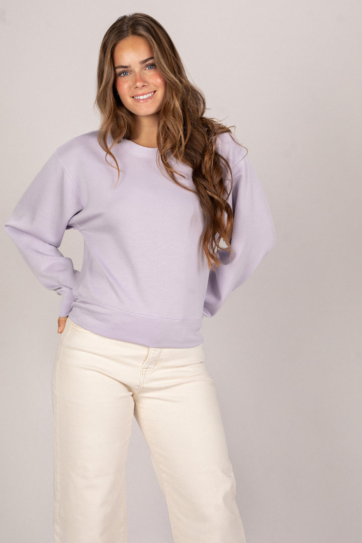 A woman with long wavy brown hair smiling while wearing a light purple relaxed-fit sweatshirt and cream-colored pants.