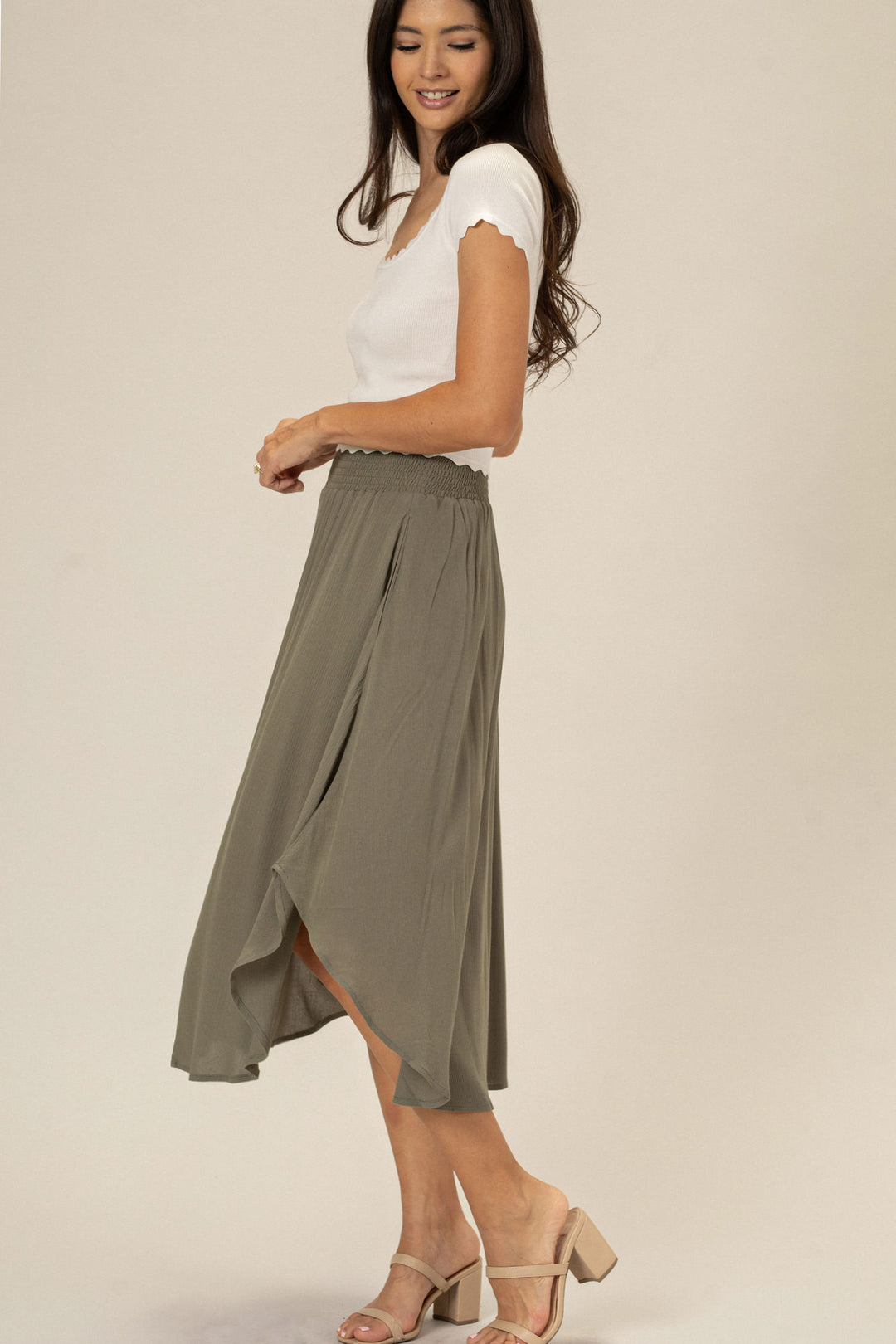 A woman wearing a flowing olive green midi skirt with an elastic waistband and a rounded hemline, paired with a white fitted top and beige heeled sandals.