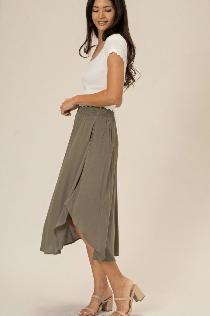 A woman wearing a flowing olive green midi skirt with an elastic waistband and a rounded hemline, paired with a white fitted top and beige heeled sandals.