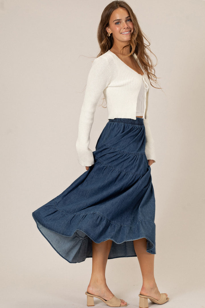 A smiling woman in a white ribbed sweater and navy blue ruffled midi skirt, posing in front of a neutral background.