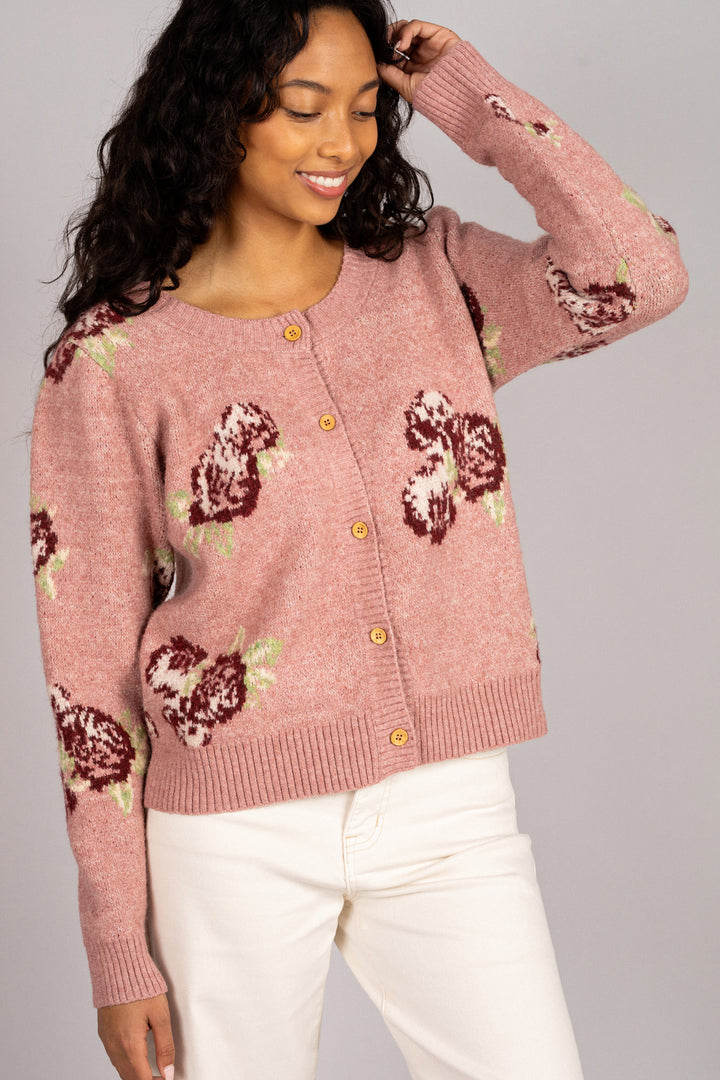 A pink knit cardigan with a floral pattern and wooden buttons, worn by a smiling woman with long, dark hair.