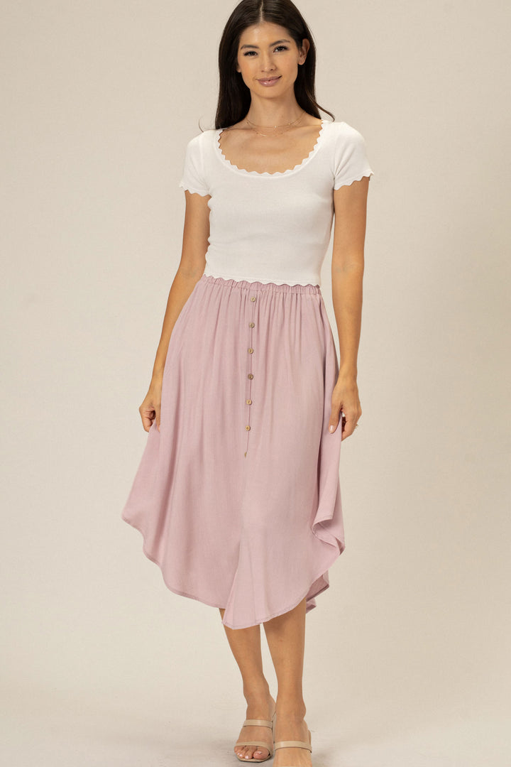 A young woman wearing a white scalloped-edge top and a mid-length, light pink button-front skirt with a ruched waistband.
