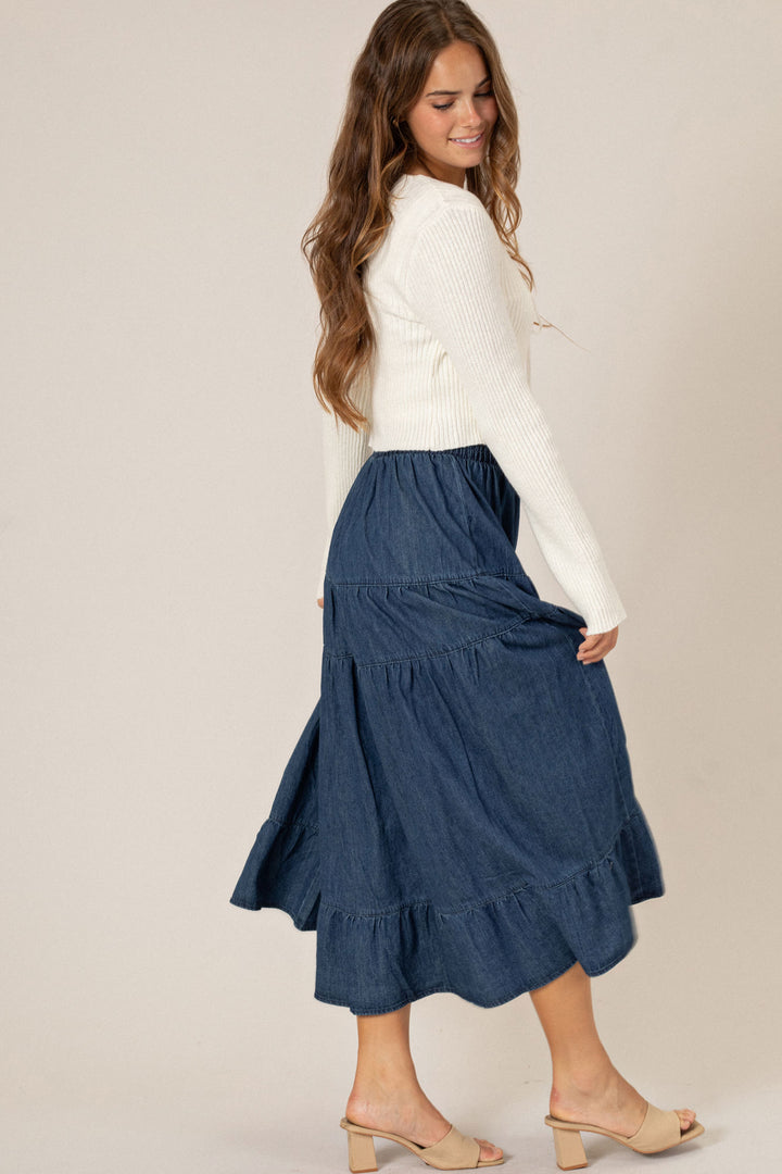 A woman in a white sweater and a long denim midi skirt with a tiered, ruffled design, standing and smiling.