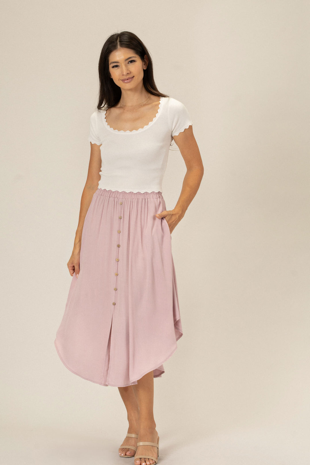 A woman wearing a white scoop-neck top and a long pink pleated skirt with buttons down the front, standing against a neutral background.