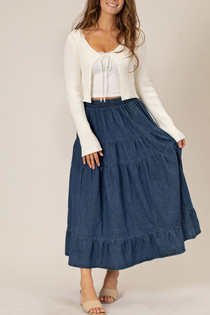 A white cropped sweater with a ribbon tie detail is paired with a flowing, mid-length denim skirt with a tiered, ruffled silhouette.