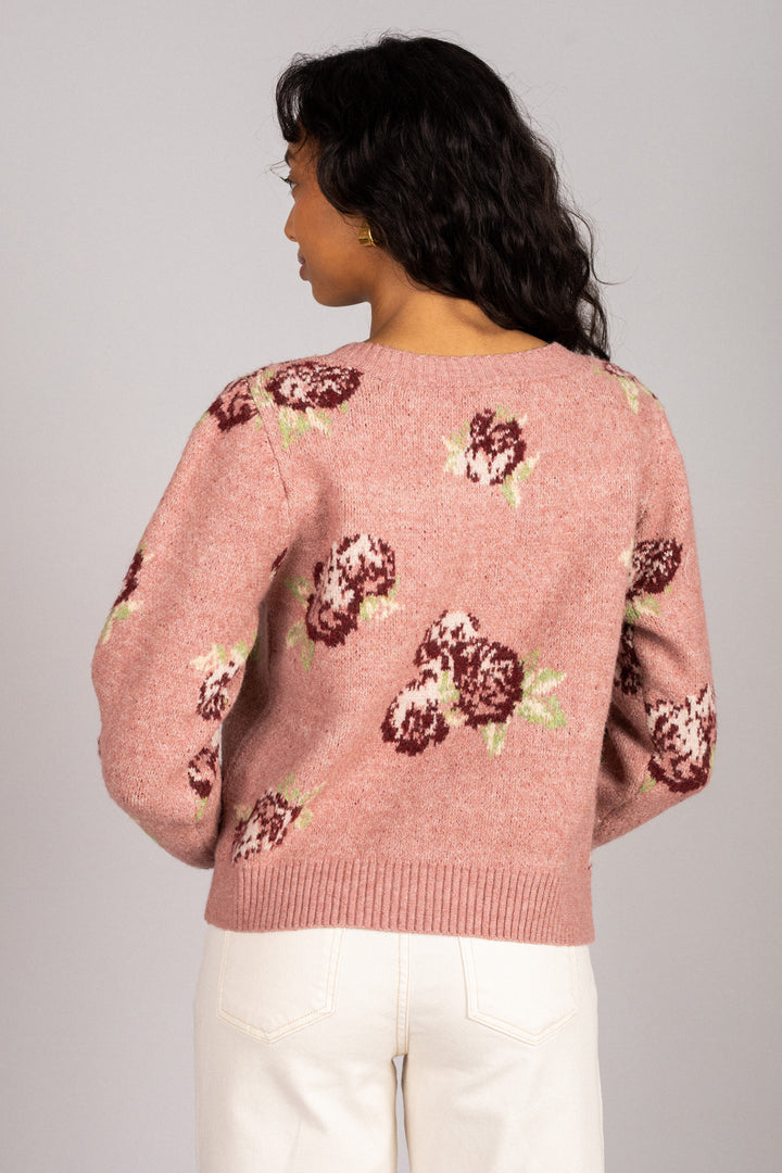 A pink knitted cardigan with floral patterns, featuring puff sleeves and a button-up closure, modeled on a woman with long dark hair.