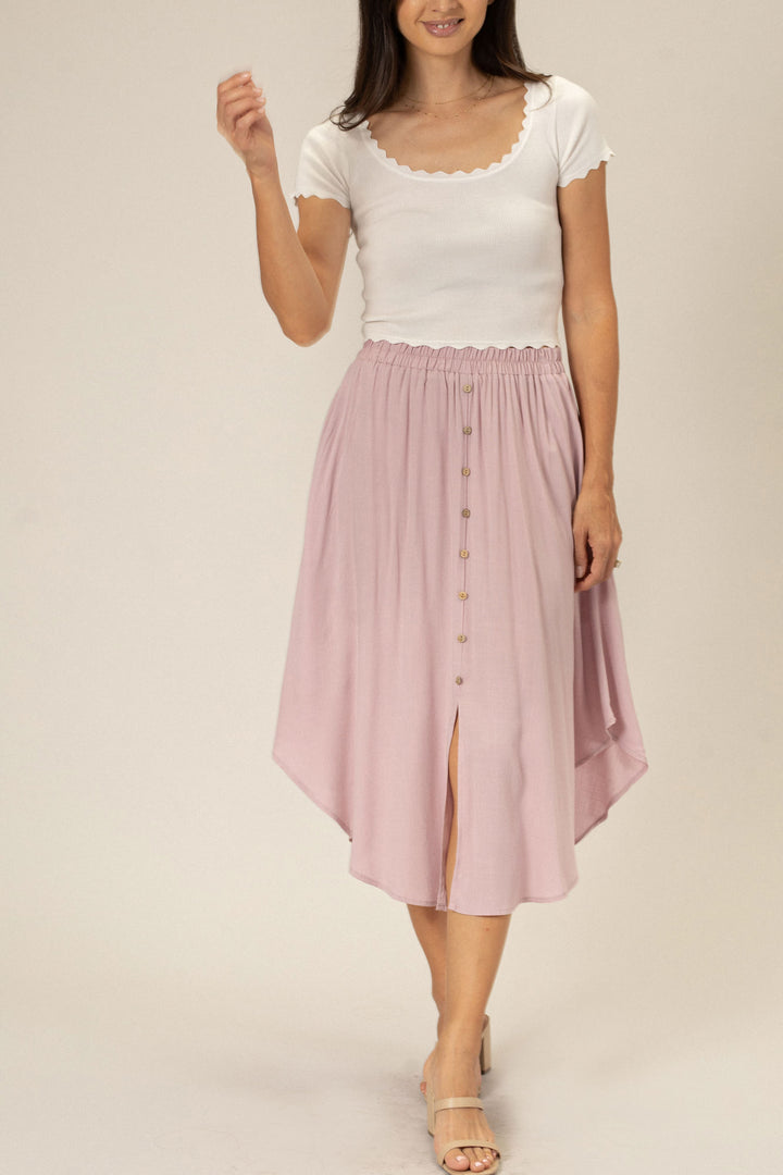 A young woman wearing a white ribbed top and a button-front dusty rose midi skirt with a flowy ruffled hem.