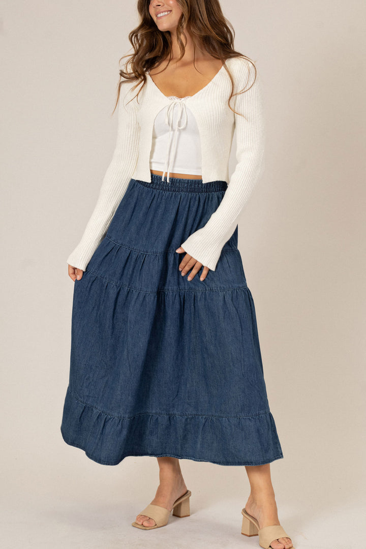 A young woman wearing a long white knit sweater with a tie front and a flowy dark denim midi skirt with tiered ruffles, paired with nude heels.