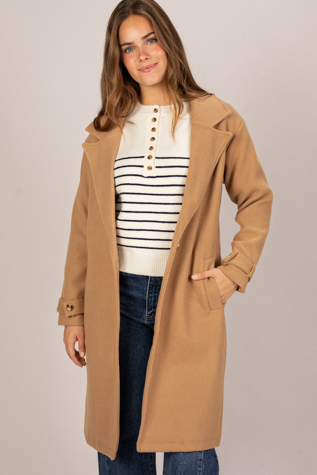 A woman wearing a beige notch-collar coat with a striped knit top, standing and smiling.