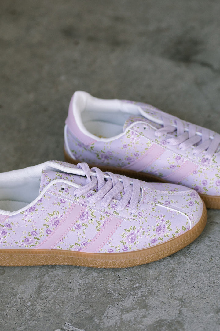 A pair of light purple sneakers featuring a delicate, all-over floral print pattern with white and yellow accents, showcasing a lace-up design and a gum rubber sole.