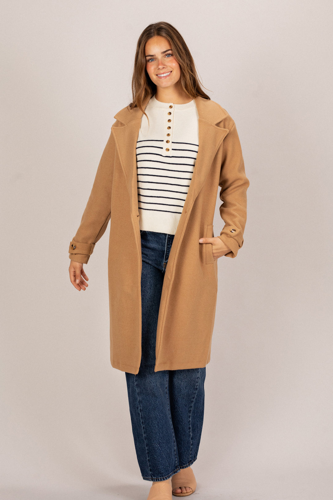 A smiling woman wearing a camel-colored notched collar coat, striped sweater, and blue jeans, modeling the Marlow Notched Collared Coat.