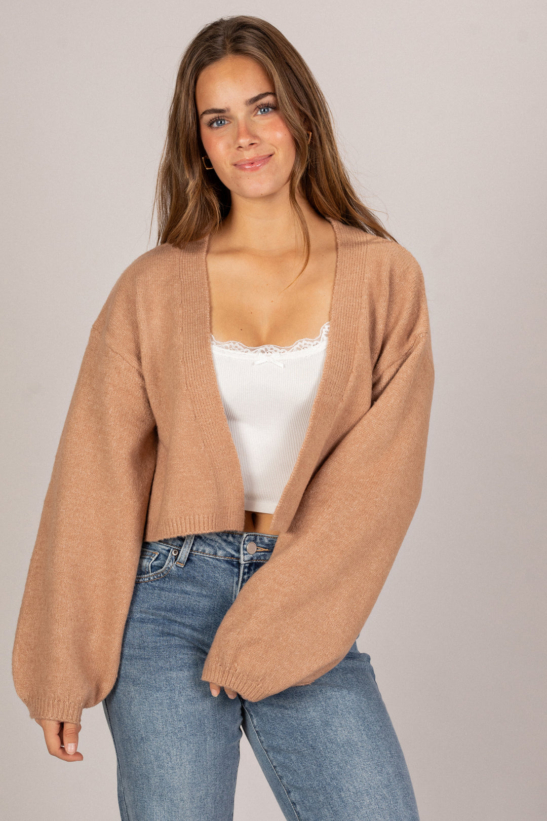 Woman wearing a brown cardigan over a white top and blue jeans on a plain background