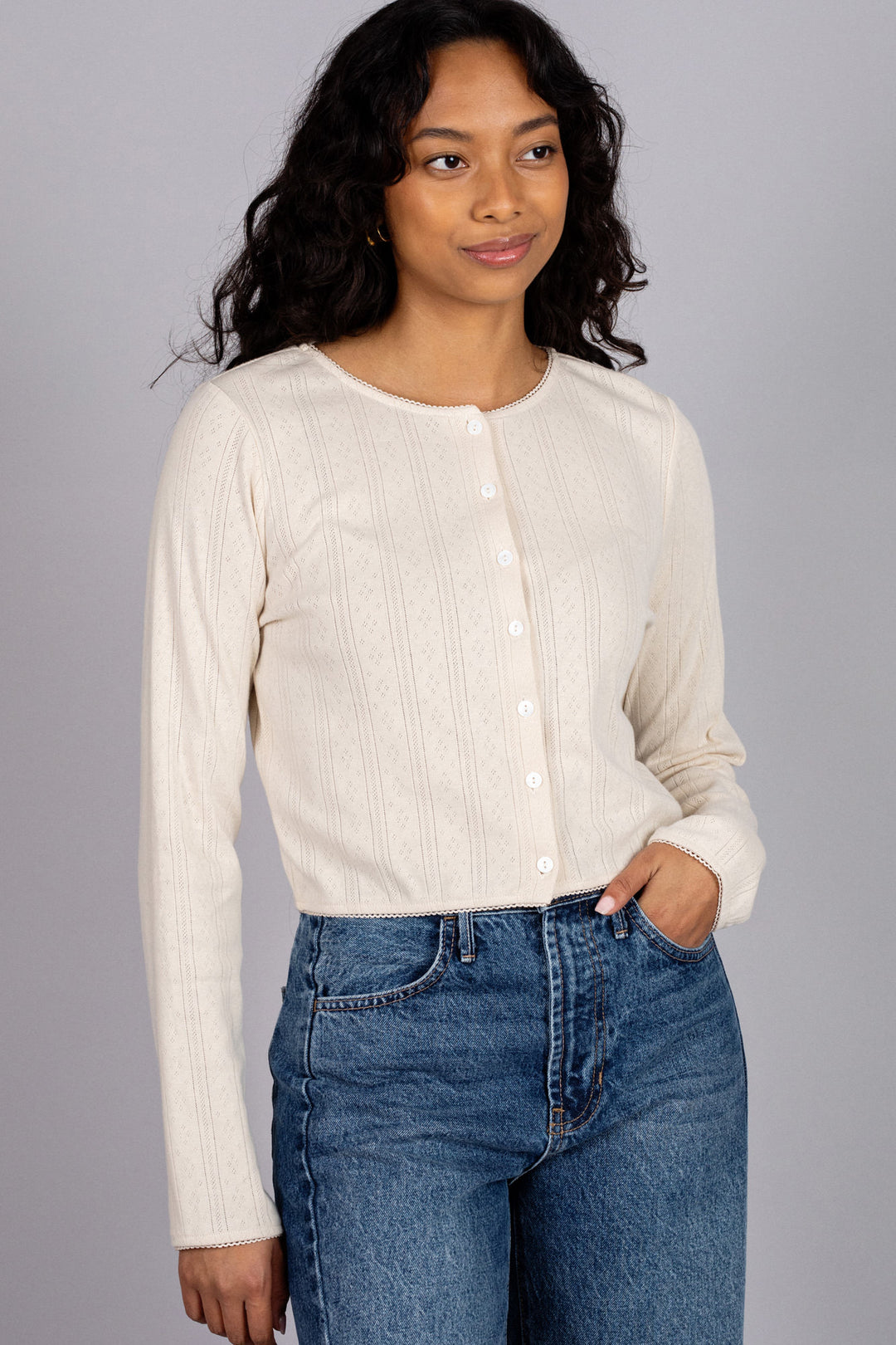A woman wearing a cream-colored, long-sleeved, buttoned top with a pointelle pattern, paired with high-waisted blue jeans.