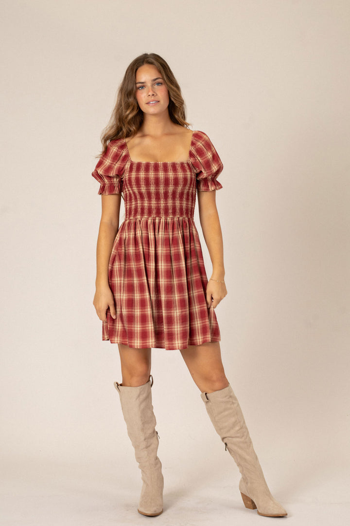 A woman wearing a red and white plaid mini dress with puffed sleeves, paired with beige knee-high boots, standing against a neutral background.
