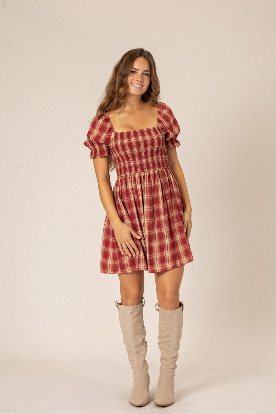 A young woman smiling and wearing a red and white plaid mini dress with puffed sleeves and a smocked bodice, paired with beige over-the-knee boots.