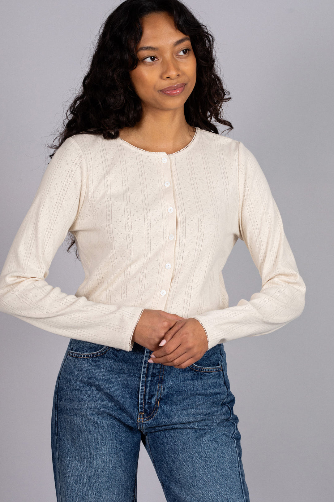 A woman in a long-sleeved, off-white buttoned sweater with a pointelle pattern, paired with high-waisted blue jeans.
