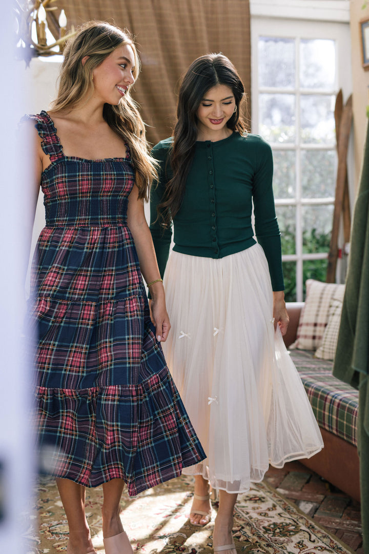 Two young women modeling dresses, one in a plaid midi dress with ruffled straps, the other in a long, white tulle skirt and green cardigan.