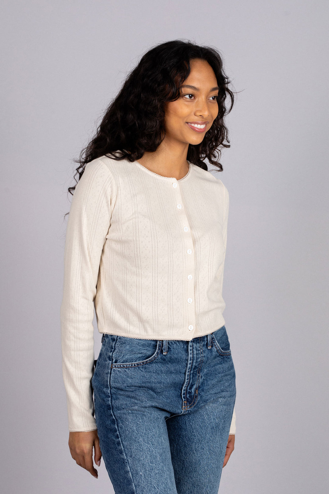 A smiling woman wearing a cream-colored, buttoned pointelle top and high-waisted blue jeans, modeling a fitted, long-sleeved knit blouse.