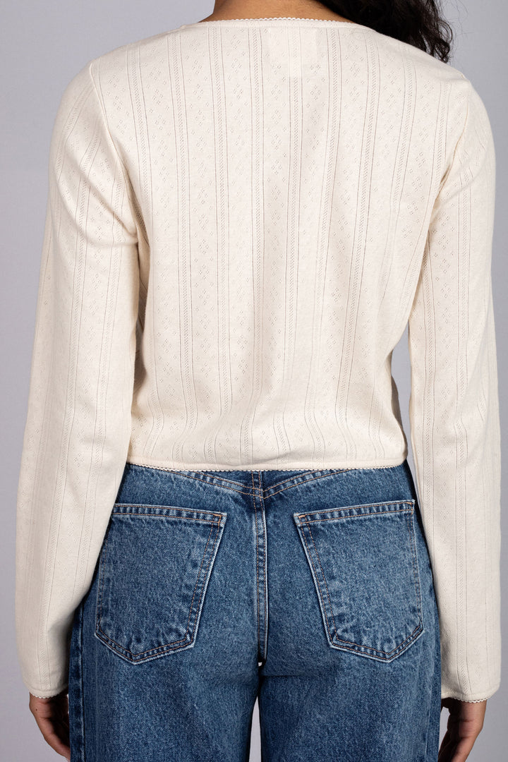 Cream-colored long-sleeved top with intricate pointelle knit patterns, worn with high-waisted denim jeans.
