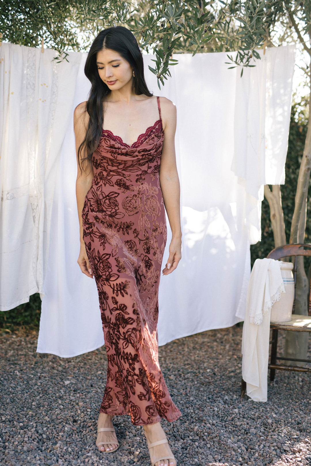 A woman wearing a burgundy floral-print maxi dress with a deep V-neck and delicate lace trim, standing against a white backdrop and olive trees.