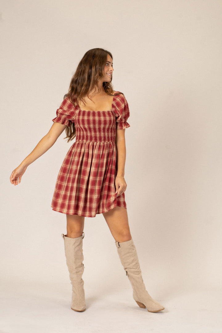 A young woman wearing a red and white plaid mini dress with puffed sleeves and a fitted waist, paired with beige knee-high boots.