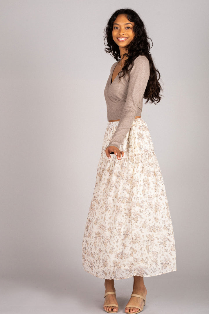 The image shows a model wearing a floral print midi skirt with a beige knit top. The skirt has a high waist and a flowing, pleated silhouette. The model is smiling and posing confidently against a plain background.