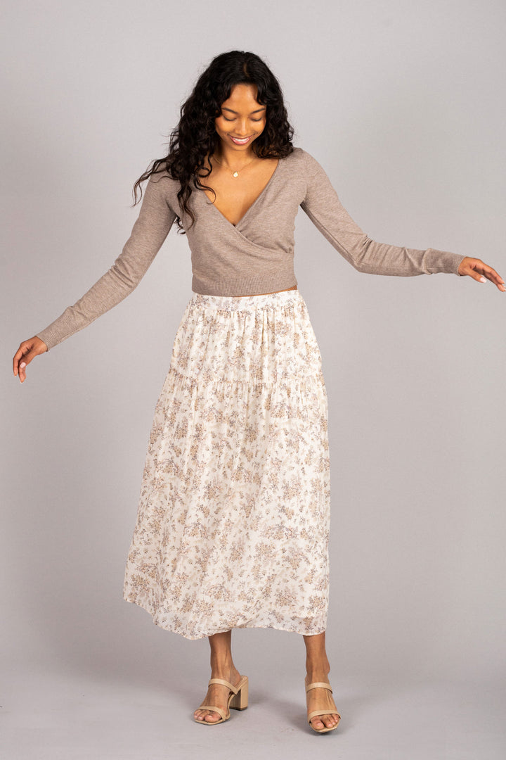 A smiling woman with long curly hair wearing a beige wrap-style sweater and a floral print midi skirt paired with nude heeled sandals.
