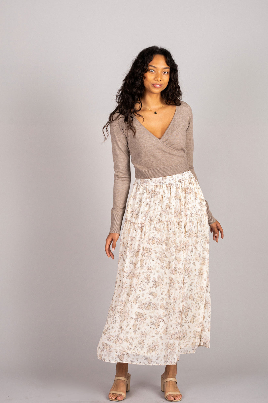 A woman with long, curly dark hair wears a fitted beige sweater and a flowy white midi skirt with a delicate floral print, paired with nude sandals.