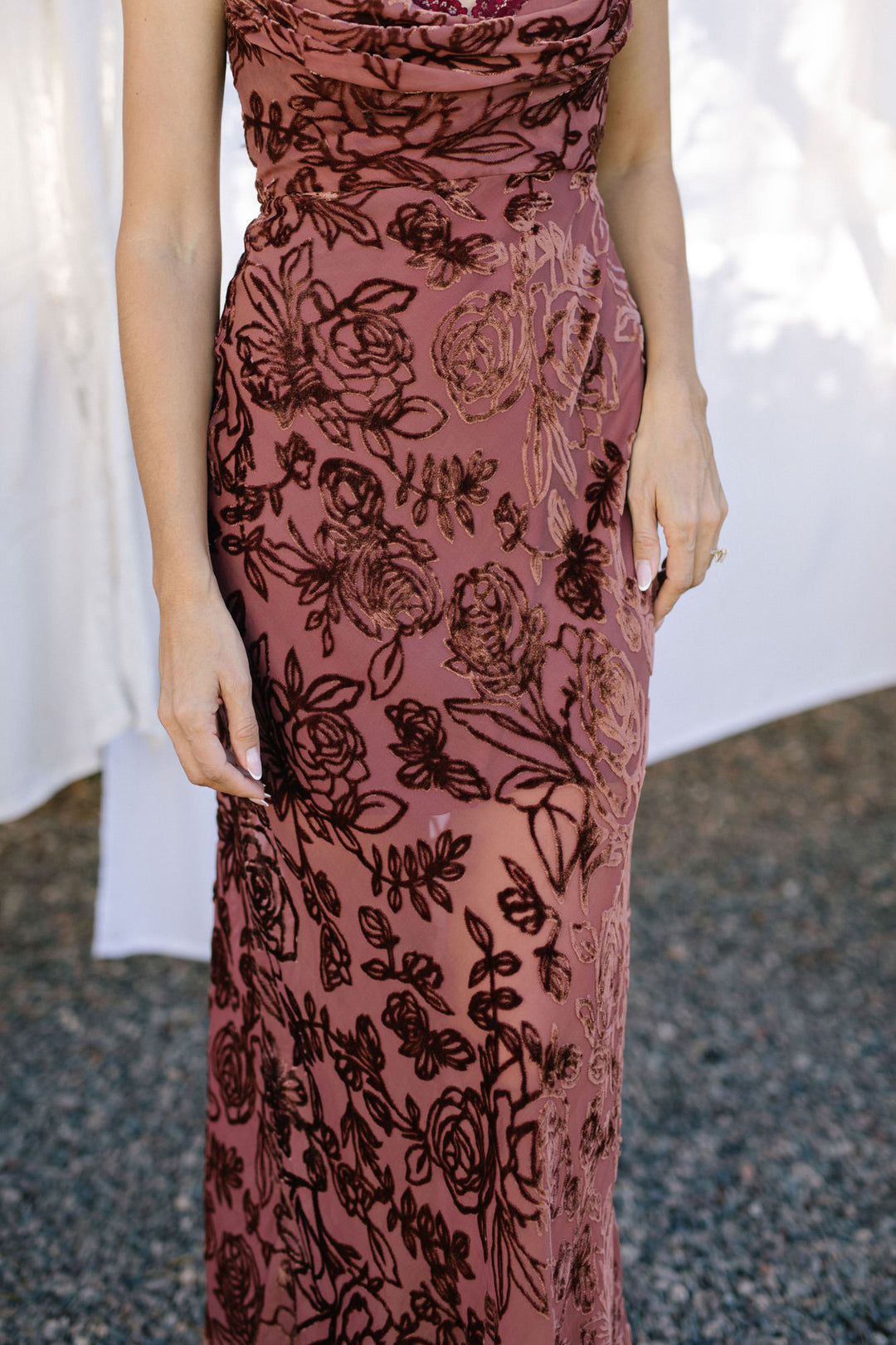 A floor-length burgundy dress with an intricate floral print and cowl neckline, featuring a form-fitting silhouette and a flowing, maxi-length skirt.