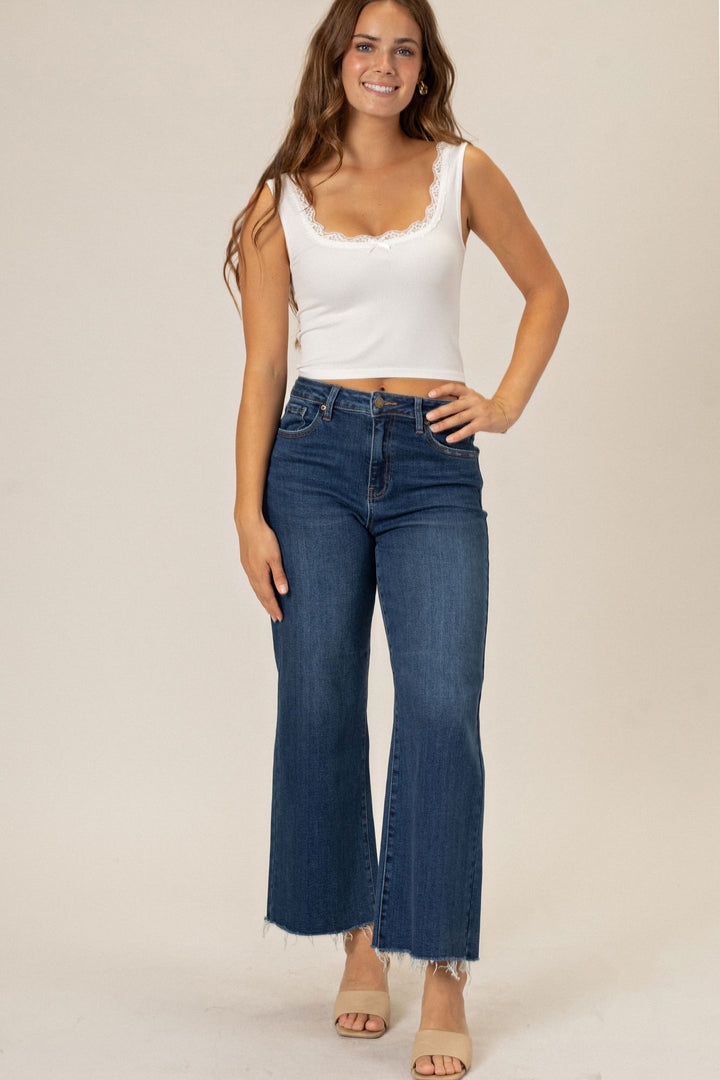 A young woman with long, wavy brown hair wearing a white lace-trimmed tank top and high-waisted dark blue wide-leg jeans with raw-edged hems, standing against a beige background.