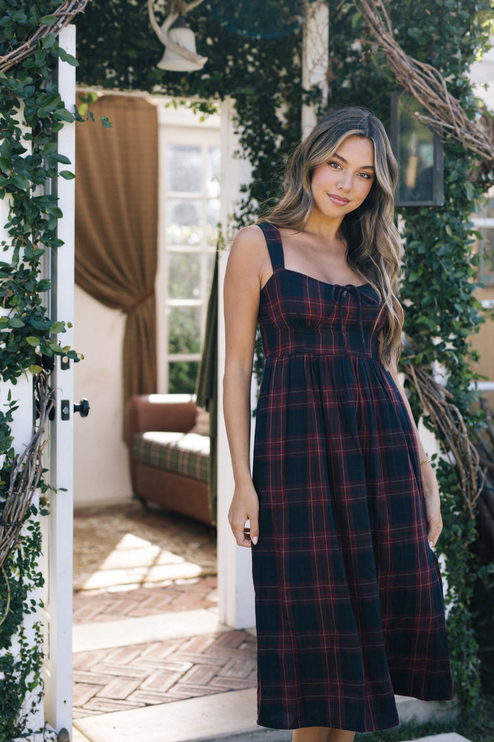 A dark plaid midi dress with a square neckline and A-line silhouette, worn by a smiling woman with long wavy hair standing in a leafy doorway.