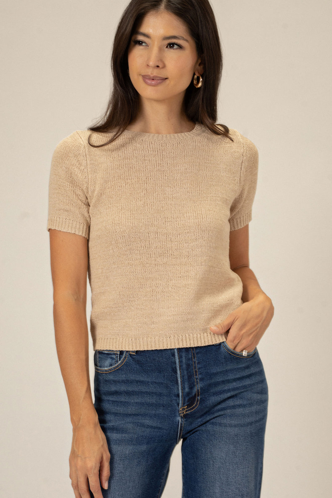 A young woman with long dark hair wearing a beige knit crew neck top and blue jeans, standing against a light background.
