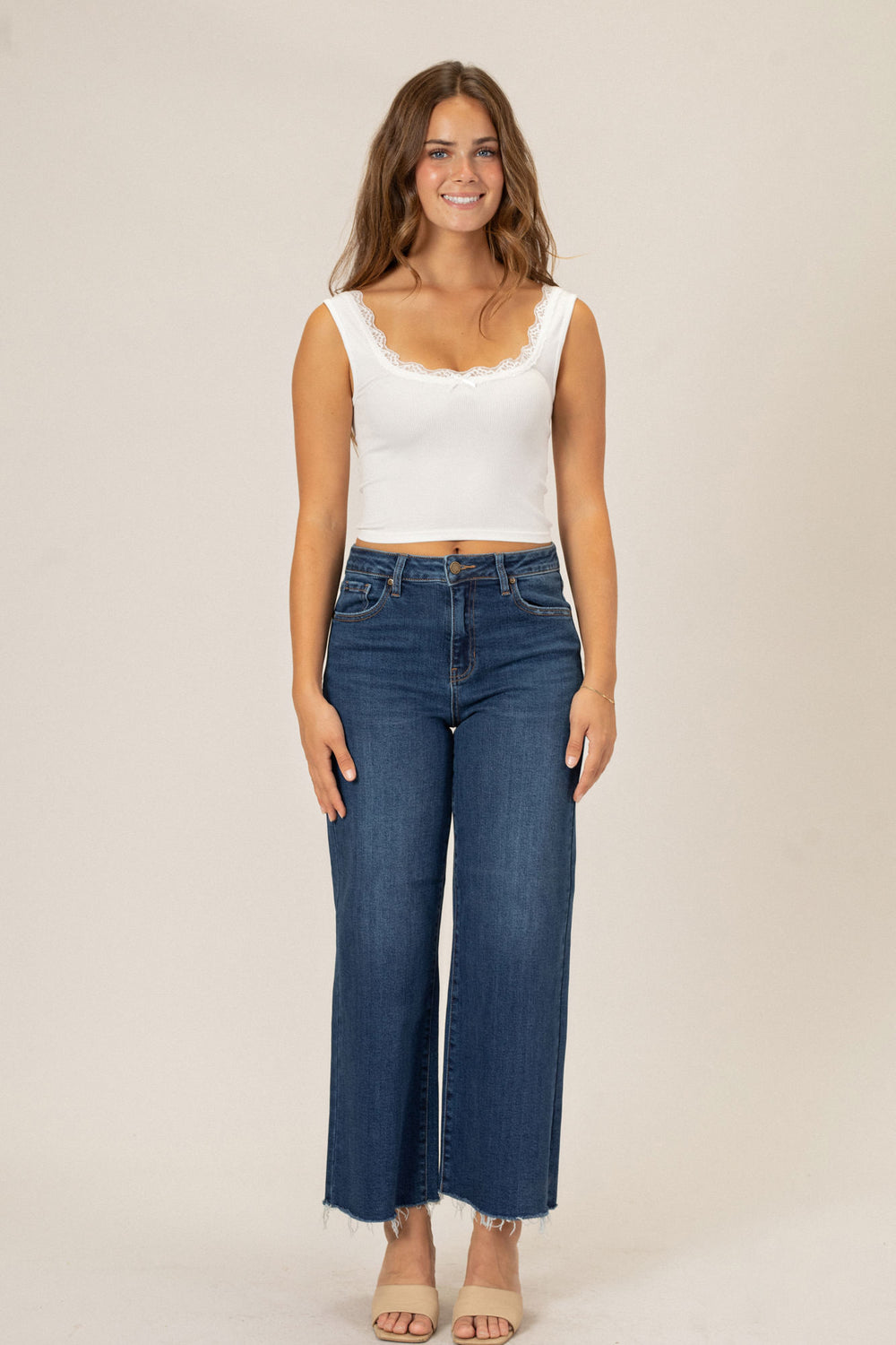 Young woman wearing a white cropped tank top and blue wide-leg denim jeans, standing against a beige background.