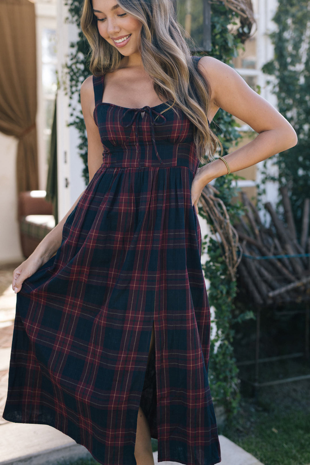A woman in a long, black and red plaid midi dress with a fitted bodice and full, flowing skirt, standing outdoors in a lush garden setting.