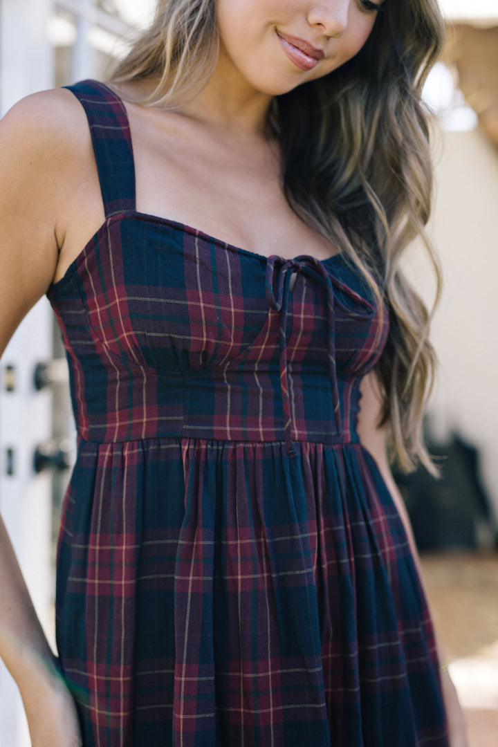 A plaid midi dress with a fitted bodice, tie detail, and flowing pleated skirt in navy, maroon, and olive tones.