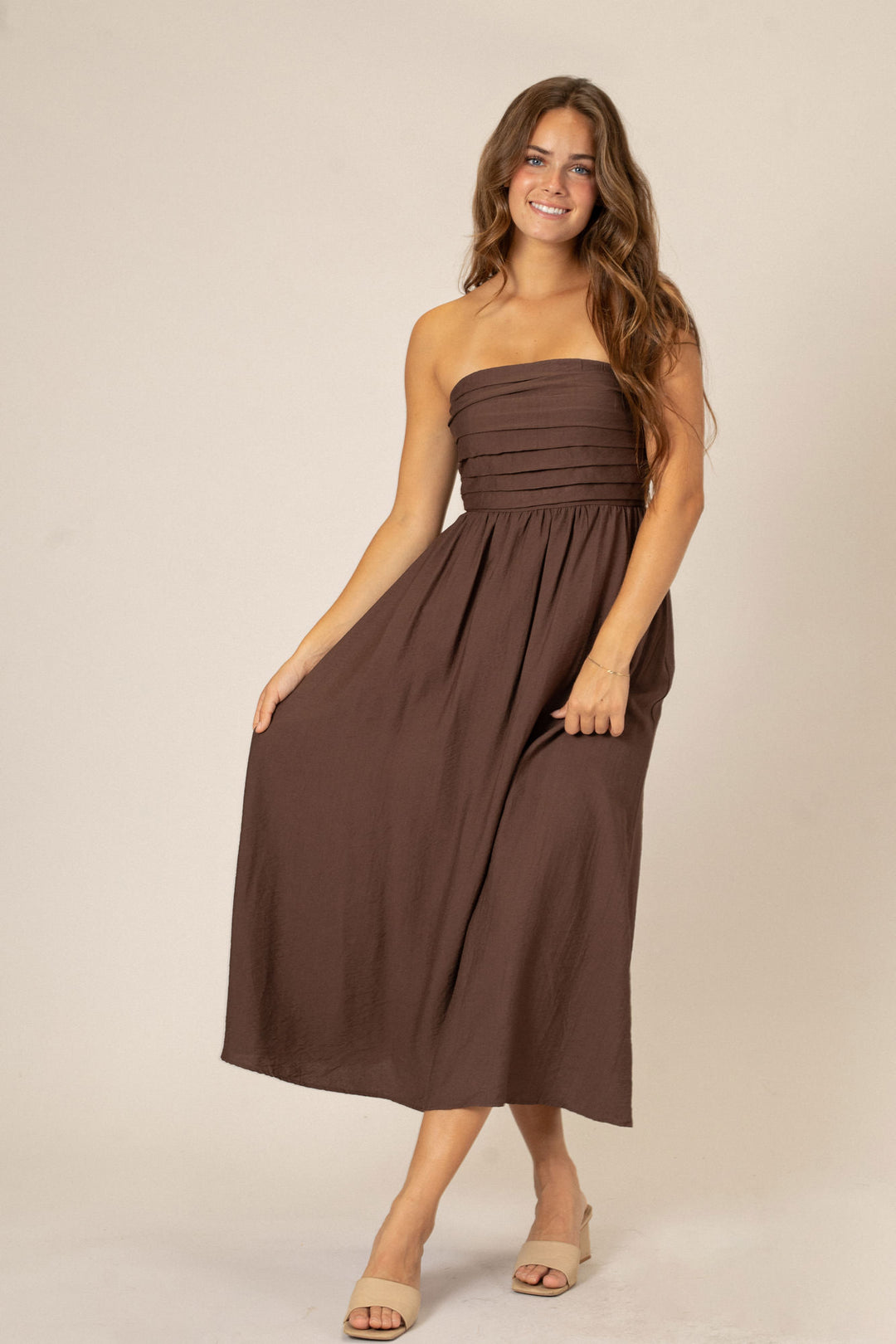 A brown strapless midi dress with pleated bodice and A-line skirt, modeled by a smiling young woman with long wavy hair.