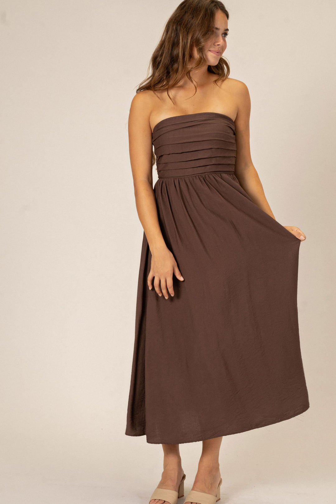 A strapless midi dress in a warm brown shade, featuring a fitted bodice with pleated detailing and a flowing, ankle-length skirt.