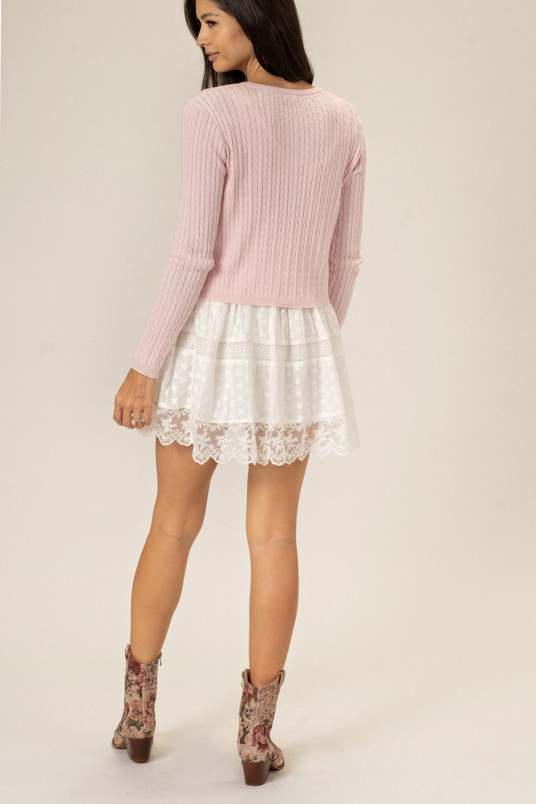 A pink cable knit sweater with a long-sleeve design, paired with a white lace skirt and floral patterned boots, modeled by a woman with long, dark hair.