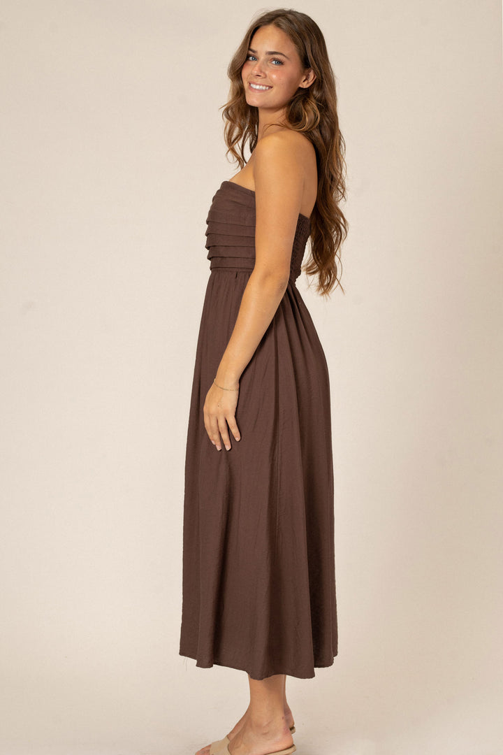 A brown strapless midi dress with pleated details and a flowy, A-line silhouette, modeled by a smiling woman with long brown hair.