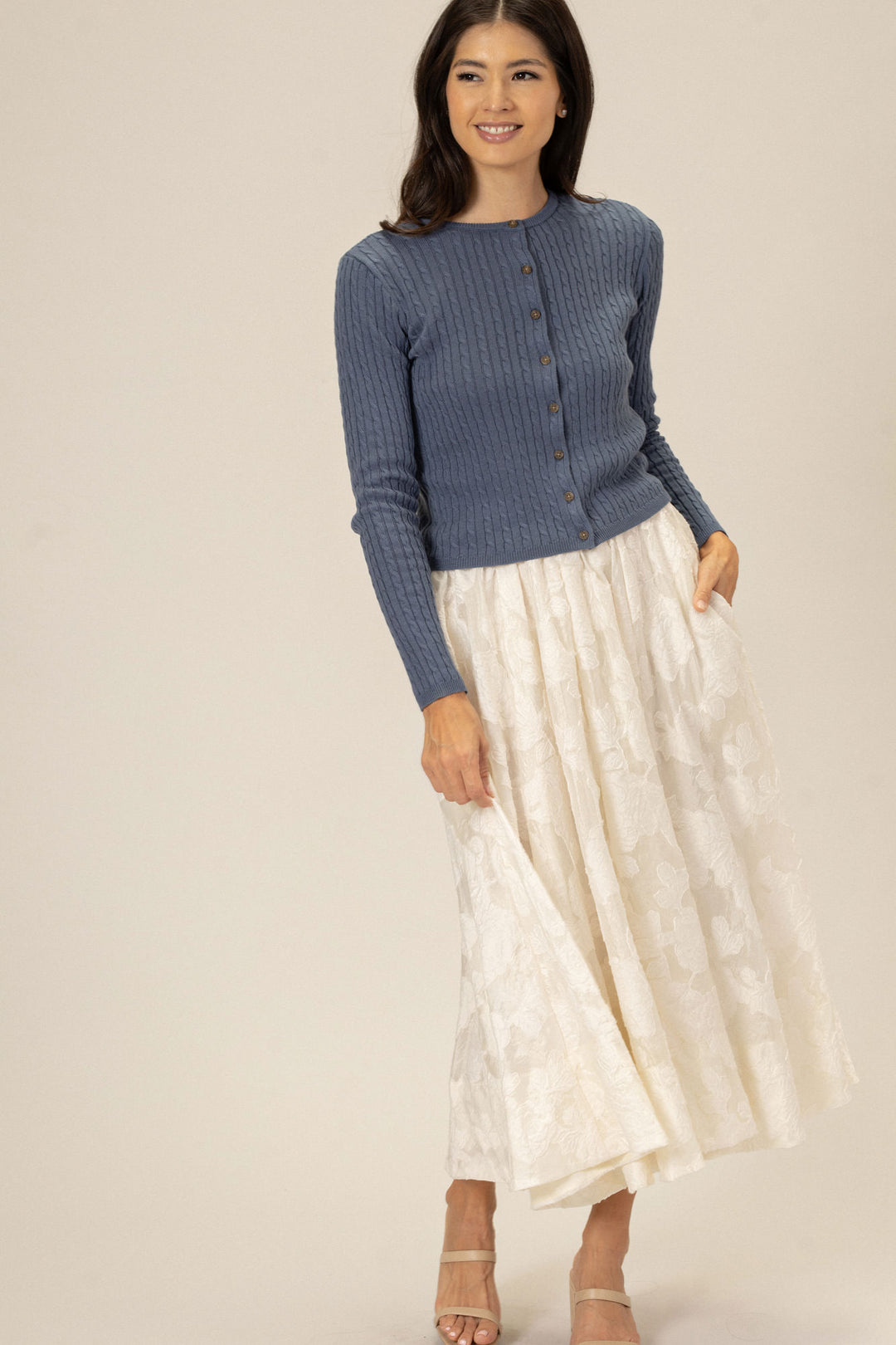 A woman wearing a gray cable-knit cardigan with wooden buttons, paired with a long, flowing white floral skirt, smiling at the camera.