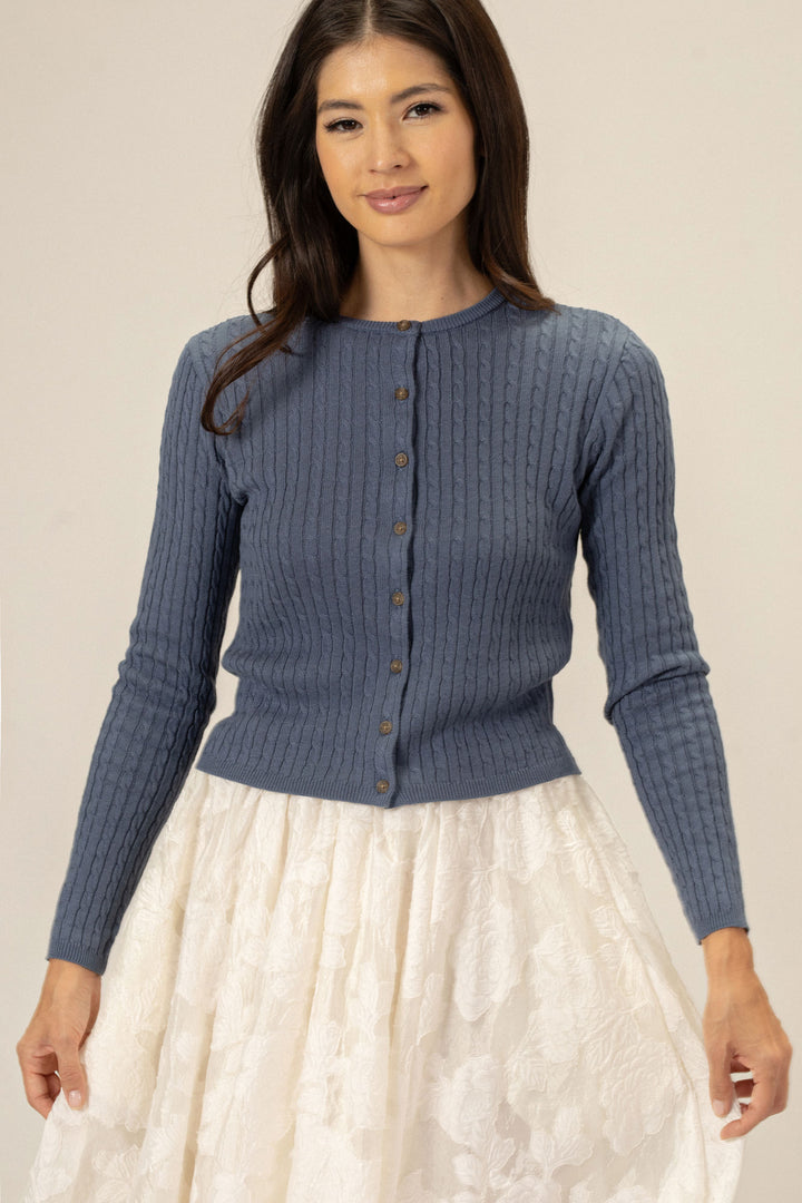 Gray cable knit cardigan with wooden buttons, paired with a white ruffled skirt, worn by a young Asian woman with long dark hair.