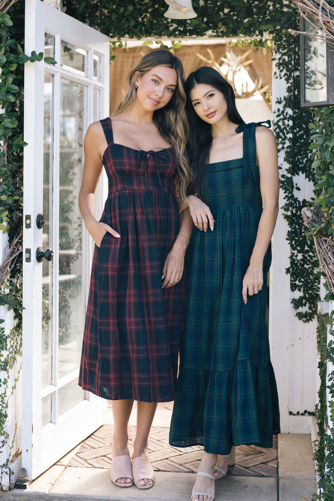 Two young women wearing plaid midi dresses, one in burgundy and one in teal, posing in a garden setting with lush greenery.